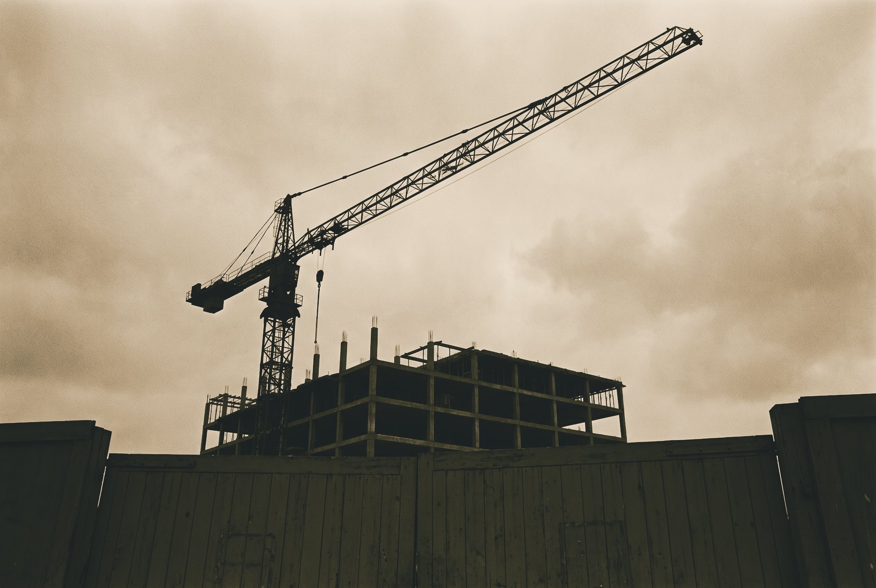 A tower crane standing still over an unfinished concrete frame, seen from a low angle behind closed site gates, with a wide empty sky.