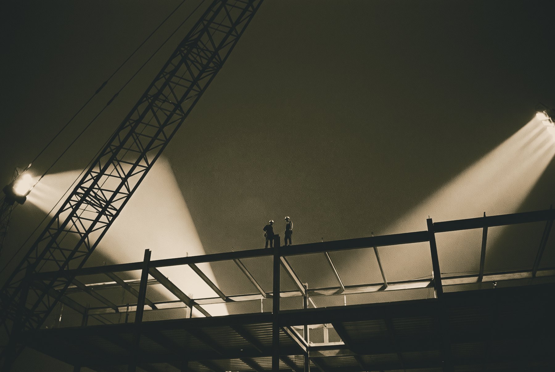 A steel building frame at night under floodlights, with a tower crane jib crossing the frame on a steep diagonal and two small silhouetted steelworkers standing on a beam.