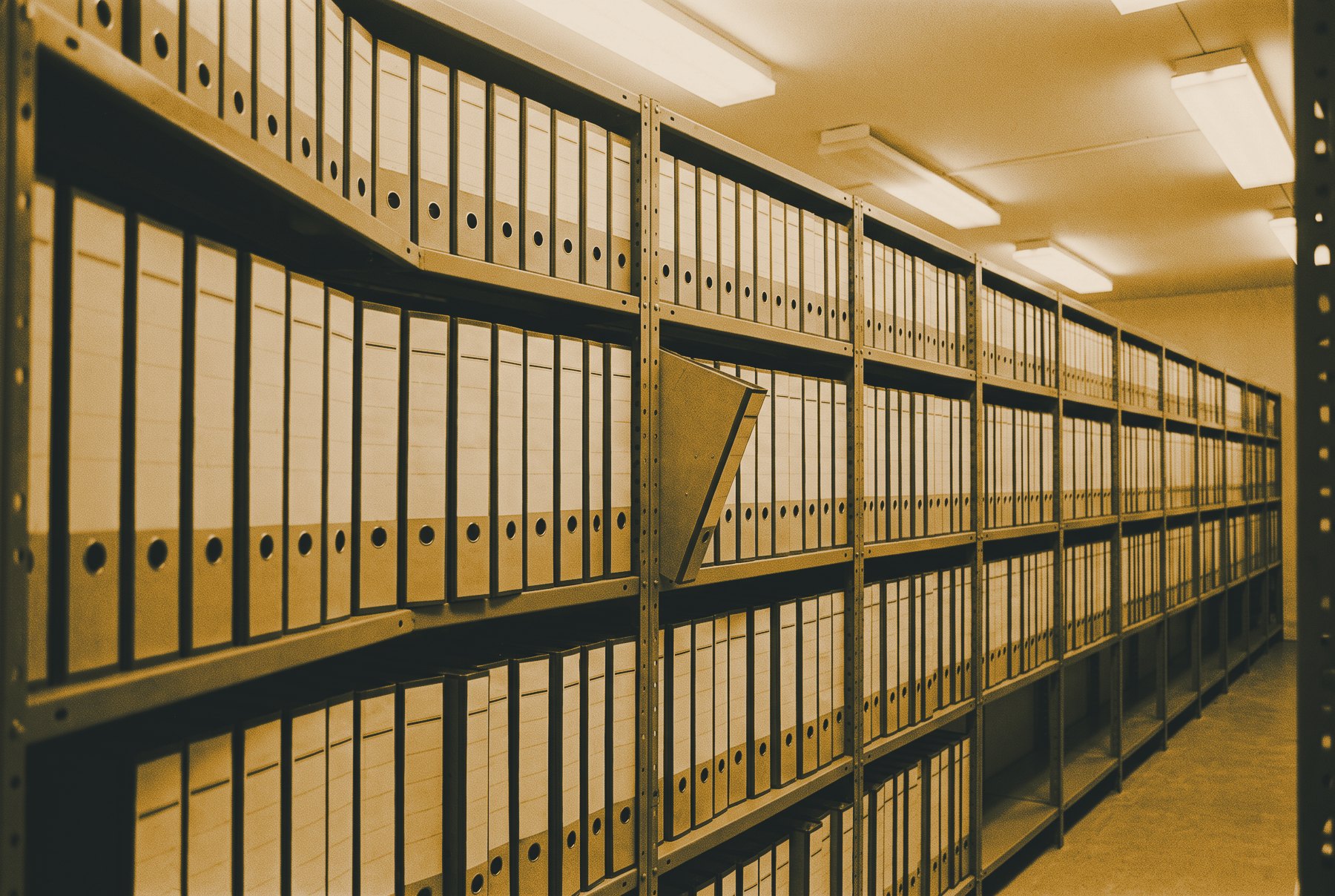 A long row of identical lever arch files with blank spines on grey steel shelving, receding to a vanishing point, with one file pulled half out.